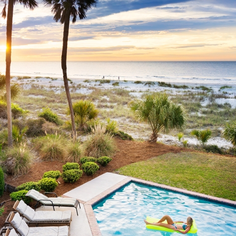 woman lounging on a pool floatie in a pool next to the beach during sunset